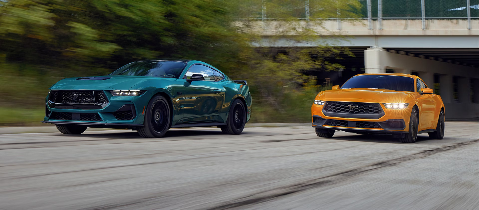 two ford mustangs driving on a road with a bridge in the background